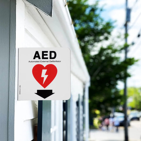 Front view of a white 6x6 inch acrylic AED sign with red heart and downward arrow mounted on a building exterior.