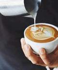 Barista pouring milk from a 20 oz stainless steel milk frothing pitcher for espresso and latte art foam into a cappuccino