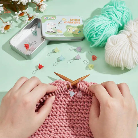 Flower Stitch Markers Set Of 18 In 9 Colors For Knitting Crochet And Weaving, shown with yarn and knitting in progress on a green table.