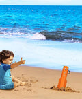 Beach scene with child, orange 3 In 1 Ultralight Backpacking Trowel Shovel And Potty Tool with ergonomic handle placed in the sand.