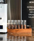 12 Piece Single Dose Coffee Bean Storage Tubes With One Way Exhaust Valve Glass Vials On Sturdy Walnut Wood Base and Easy Pouring Funnel displayed on counter.