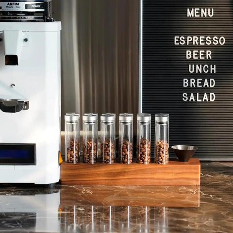 12 Piece Single Dose Coffee Bean Storage Tubes With One Way Exhaust Valve Glass Vials On Sturdy Walnut Wood Base and Easy Pouring Funnel displayed on counter.