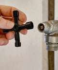 Hand holding 4-Way Universal Utility Cabinet Key 2 Pack In Black For Gas Meters, Water Panels, And Electric Boxes near a valve