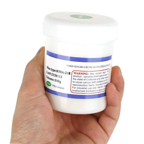 Hand holding a jar of RMA 218 Flux Paste For BGA Reballing And Soldering, 100g size, white container.