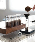 12 Piece Single Dose Coffee Bean Storage Tubes With One Way Exhaust Valve Glass Vials On Sturdy Walnut Wood Base and Easy Pouring Funnel in use.