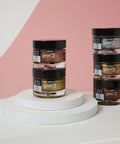 Pebeo gilding wax jars in copper, bronze, gold, and silver on a white pedestal against a pink backdrop.
