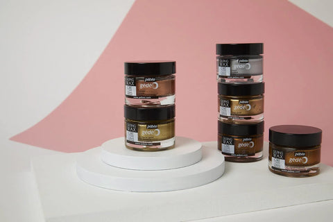 Pebeo gilding wax jars in copper, bronze, gold, and silver on a white pedestal against a pink backdrop.