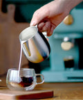 Hand pouring milk from a stainless steel pitcher into a glass cup next to an Electric Coffee Maker With Auto Milk Frother For Smooth Cappuccino Sips Transparent Water Reservoir Safety Valve Removable Drip Tray Stainless Steel Frame Smart Controls A Game Changer For Weekend Coffee Crews.