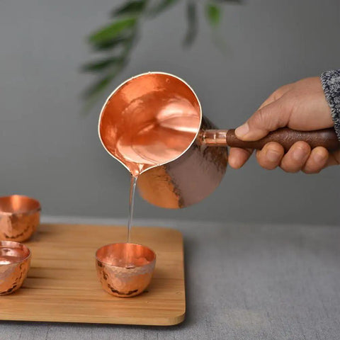 Pouring from Copper Teapot With Wooden Handle Dope Hammered Turkish Coffee Pot Multipurpose Kettle For Tea Coffee Maple Syrup And Sauce Heating Hand Crafted In Turkey For Your Home Kitchen into cups on a wooden board