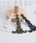 Three metal decorative hinges in white, bronze, and black finishes displayed on a wooden surface.