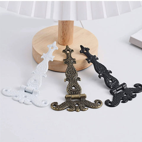 Three metal decorative hinges in white, bronze, and black finishes displayed on a wooden surface.