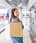 Person holding a brown paper bag with handles in a shopping mall, featuring Bulk 500 Pack Heavy Duty Kraft Paper Bags 10x13 Inches With Handles 10x5x13 Inches Brown Gift Shopping Retail Bags Recycled Paper Bags For Parties And Gifts.
