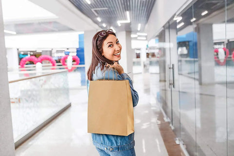 Person holding a brown paper bag with handles in a shopping mall, featuring Bulk 500 Pack Heavy Duty Kraft Paper Bags 10x13 Inches With Handles 10x5x13 Inches Brown Gift Shopping Retail Bags Recycled Paper Bags For Parties And Gifts.