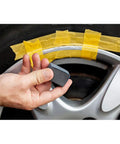 Hand holding a block near a black rim with tape, demonstrating repair with Solid Alloy Wheel Repair Kit For Black Rims Smooths Out Curb Rash And Road Scuffs DIY Two Part Liquid Aluminum Compound Plus Spot On Black Touch Up Paint For A Fresh New Ride Glow Up.