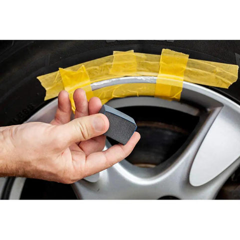Hand holding a block near a black rim with tape, demonstrating repair with Solid Alloy Wheel Repair Kit For Black Rims Smooths Out Curb Rash And Road Scuffs DIY Two Part Liquid Aluminum Compound Plus Spot On Black Touch Up Paint For A Fresh New Ride Glow Up.