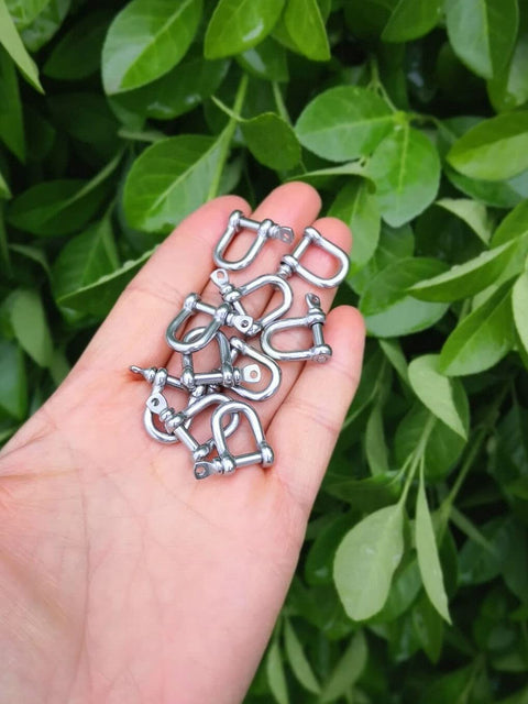 Hand holding 316 Stainless Steel D Shackle Mini Shackles For Paracord Jewelry And Bracelet Hardware Built Tough Marine Grade Rigging 1 Eighth Inch Pin Opening Durable Pack Of 10 For Survival Gear And Craft Projects against a leafy background