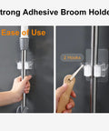 Two Pack Adhesive Broom And Mop Holder With Rubber Grips, Wall Mount Organizer With Hooks mounted on wall with hands holding handle