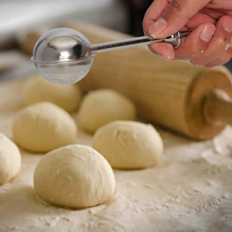 One-handed operation stainless steel flour duster sifter shaking powdered sugar over dough with rolling pin in the background.