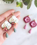 Hand holding floral polymer clay earrings with teardrop details, pink and green tones, displayed on a neutral background.
