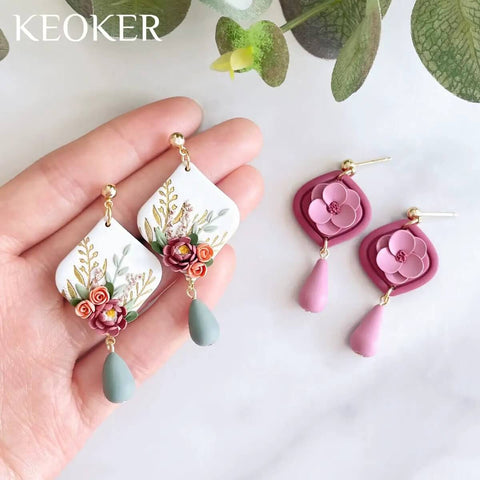 Hand holding floral polymer clay earrings with teardrop details, pink and green tones, displayed on a neutral background.