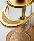 Top Shelf Belgian Siphon Coffee Maker With Stunning Gold Finish Built From Heavy Glass And Steel Frame Relies On Steam Heat And Gravity Action To Pull Silky Smooth Brews Fast Sick Flex For True Coffee Geeks Wanting Elite Taste At Home Or Work - close-up view of gold handle.
