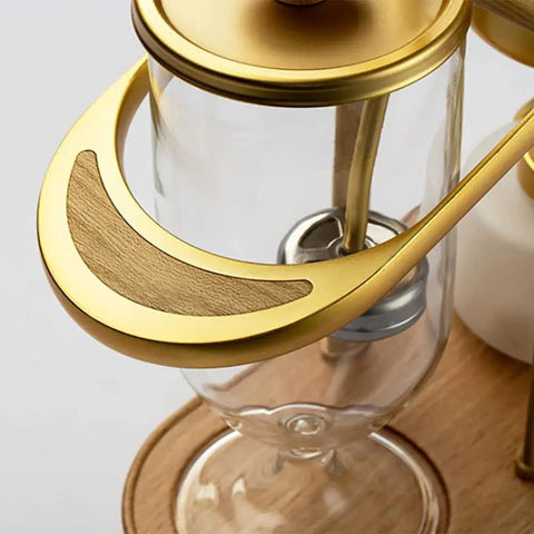 Top Shelf Belgian Siphon Coffee Maker With Stunning Gold Finish Built From Heavy Glass And Steel Frame Relies On Steam Heat And Gravity Action To Pull Silky Smooth Brews Fast Sick Flex For True Coffee Geeks Wanting Elite Taste At Home Or Work - close-up view of gold handle.