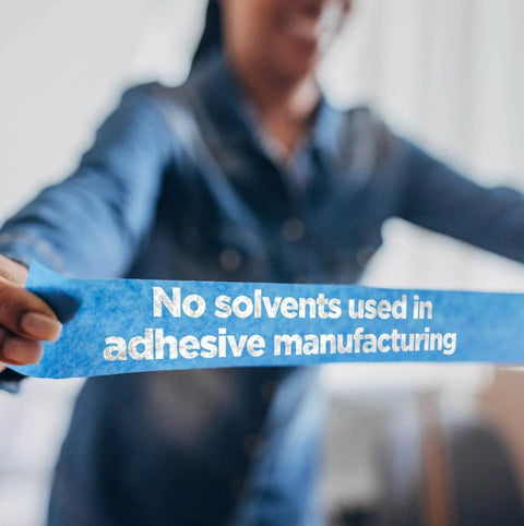 Person holding blue painter's tape with text 'No solvents used in adhesive manufacturing' visible