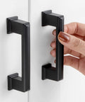 Hand holding 10 Pack 3 Inch Hole Center Matte Black Cabinet Pulls And Handles For Cabinets Drawers Dressers Vanities Closets Durable Metal Hardware With Included 1 Inch And 1 And A Half Inch Screws For Easy Installation on white cabinet