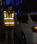 Person wearing Hi Vis Safety Vest With Multi Pockets 5 Pack Reflective Gear For Work Construction Parking Warehouse next to a car at night.
