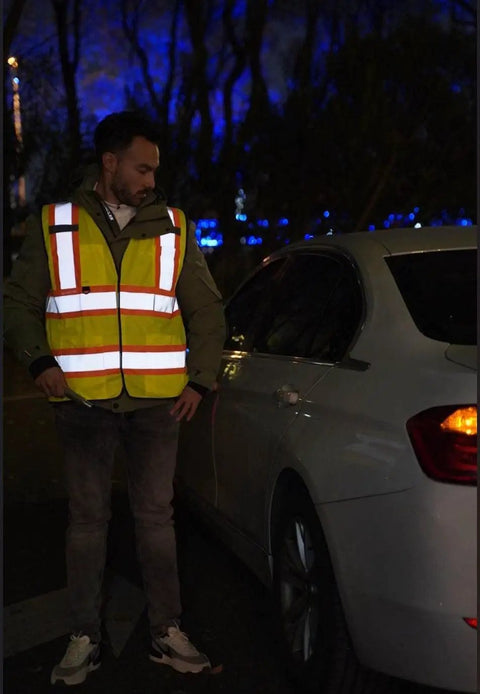 Person wearing Hi Vis Safety Vest With Multi Pockets 5 Pack Reflective Gear For Work Construction Parking Warehouse next to a car at night.