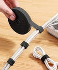 Hand holding black roll of Six Point Five Foot Two Roll Adjustable Cable Ties Reusable Hook and Loop Fastening for Cable Organizing and Wire Management Black Ideal for Home and Office Cable Management Quick Install Reusable and Adjustable to Your Needs, wrapped around power cords on a wooden desk.