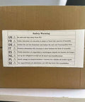 Safety warning label on a cardboard box in multiple languages, cautioning to stay away from fire, photographed on a desk.