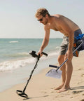 Man using heavy duty stainless steel sand scoop for metal detecting with long handle on beach, part of treasure hunting kit.