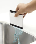 Hand using Kitchen Sink Squeegee And Countertop Brush Must Have For Wet And Dry Spills Quick Cleanup Dishwasher Safe White Silicone Squeegee Nylon Bristles Hang From Counter Or Sink Edge to wipe liquid into sink