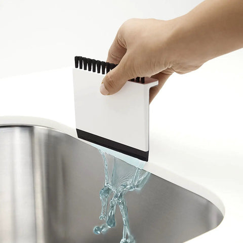 Hand using Kitchen Sink Squeegee And Countertop Brush Must Have For Wet And Dry Spills Quick Cleanup Dishwasher Safe White Silicone Squeegee Nylon Bristles Hang From Counter Or Sink Edge to wipe liquid into sink