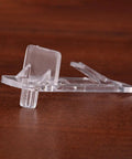 Close-up of a clear plastic shelf support clip with locking pin on a wooden surface, part of 30 Pack Plastic Shelf Support Clips Locking Pins For Cabinets Furniture Shelving Brackets Transparent Reusable Pegs For Kitchen Bookcase Garage Storage.