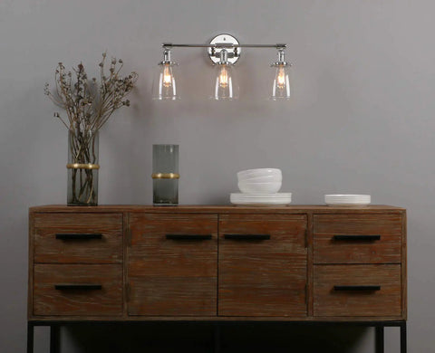 Three Light Industrial Wall Sconce With On Off Switch And Clear Glass Shade For A Cool Retro Vibe Vintage Antique Brass Vanity Lighting For Bathroom And Living Room Dimmable When Paired With A Compatible Dimmer Bell Shade Finish, installed above a wooden sideboard.