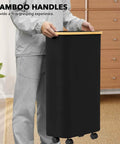 Person holding a black Slim Laundry Hamper With Wheels And Lid, 62L Rolling Clothes Basket with a bamboo handle, narrow design.