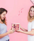 Two women exchanging a pink gift box with ribbon on a pink background, showing joy and friendship