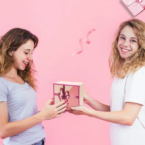 Two women exchanging a pink gift box with ribbon on a pink background, showing joy and friendship
