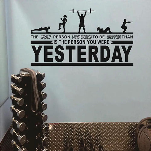 Motivational Gym Wall Decal For Home Gym Crossfit Quote Vinyl Sticker 18 By 32 Inches Black Waterproof Non Toxic Removable Decor That Inspires You To Be Better Than You Were Yesterday on a grey wall with dumbbells nearby.