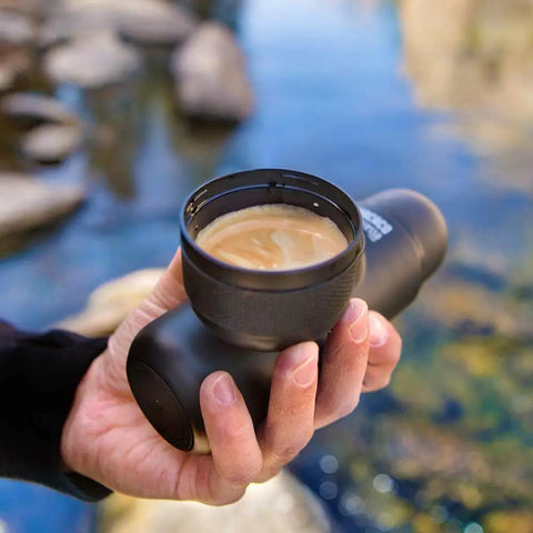 A person holding a portable hand powered espresso maker with manual piston action, near a river, for travel and office brew use.