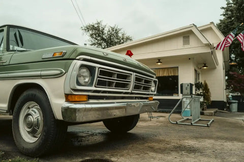 Vintage green and white truck in front of a retro gas station with American flags on a cloudy day.