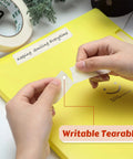 Hands tearing a piece of beige tape over a yellow notebook, showcasing the easy tear feature of the 30 rolls masking tape bulk 55 yards each for classroom labeling crafts office school projects super handy easy tear and clean removal beige white 1 inch general purpose tape.