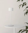 Super Sleek Portable LED Lamp In Warm Gray on round white table with green plant background. Ideal for home or desk use.