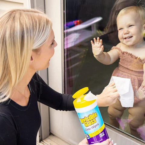 Invisible Glass Glass Wipes bottle with yellow cap; woman cleaning a glass door as a baby watches outside