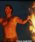A person triumphantly stands by a large campfire at night, arms raised and smiling, with subtitles saying 'I have made fire!'
