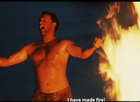 A person triumphantly stands by a large campfire at night, arms raised and smiling, with subtitles saying 'I have made fire!'