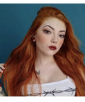 Portrait of a woman with long orange hair, wearing a white top, showcasing a nose stud and piercings against a blue background.