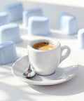 White ceramic espresso cup with saucer and spoon on a white surface; blue sugar cubes blurred in background.